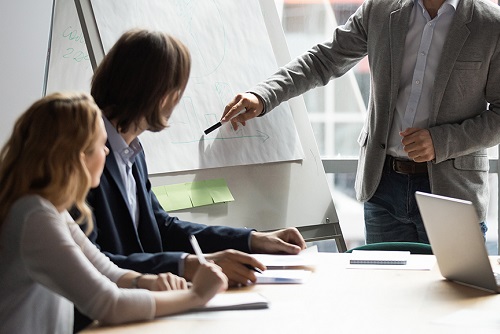 Skilled 30s caucasian leader in eyeglasses coaching interested business people, giving educational workshop presentation in office. Concentrated employees listening to professional coach at workshop.